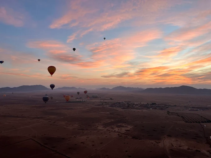 Hot air balloons floating over Moroccan desert landscape at sunrise with dramatic pink and orange sky, mountains in distance, showcasing scenic aerial adventures available in vacation packages to Marrakech