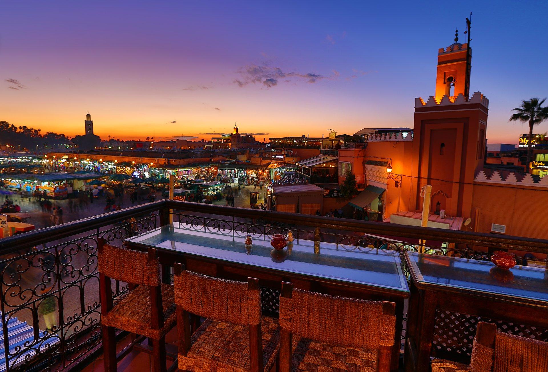 Panoramic view from a riad rooftop restaurant in Marrakech showing the bustling Jemaa el-Fnaa square and historic minaret at twilight, highlighting the magical ambiance available through vacation packages to Marrakech