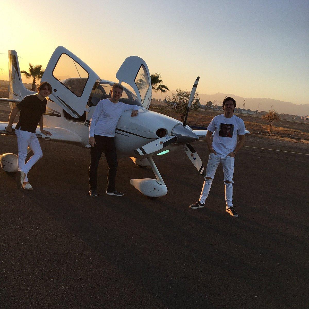 Three tourists posing with small private aircraft at sunset airfield in Morocco with palm trees and mountains in background, showcasing luxury travel experiences included in vacation packages to Marrakech