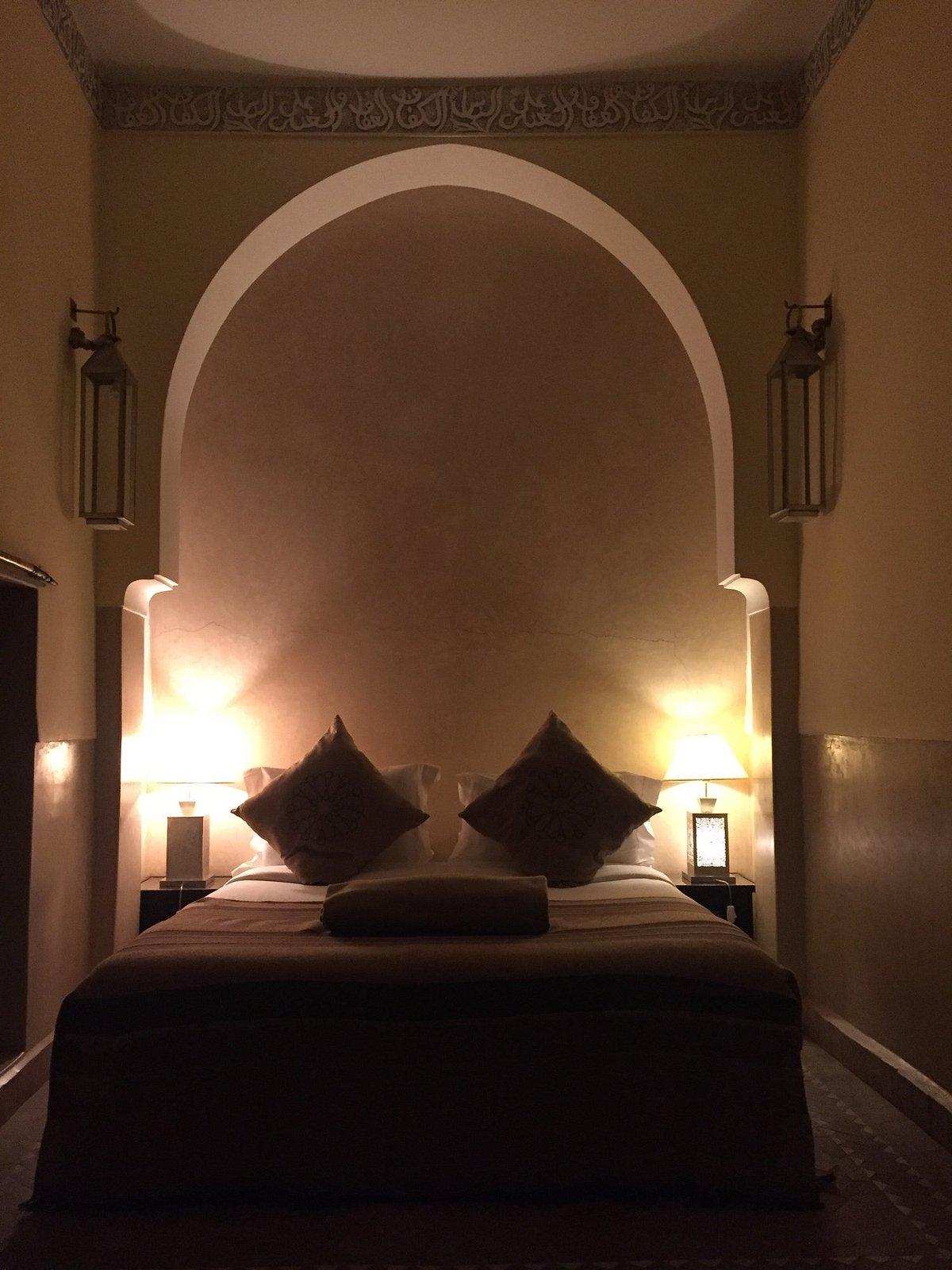 Elegant hotel bedroom with traditional Moroccan architecture featuring ornate Islamic calligraphy ceiling borders, horseshoe arch alcove, ambient lighting, and luxurious bedding at one of the best resorts in Morocco