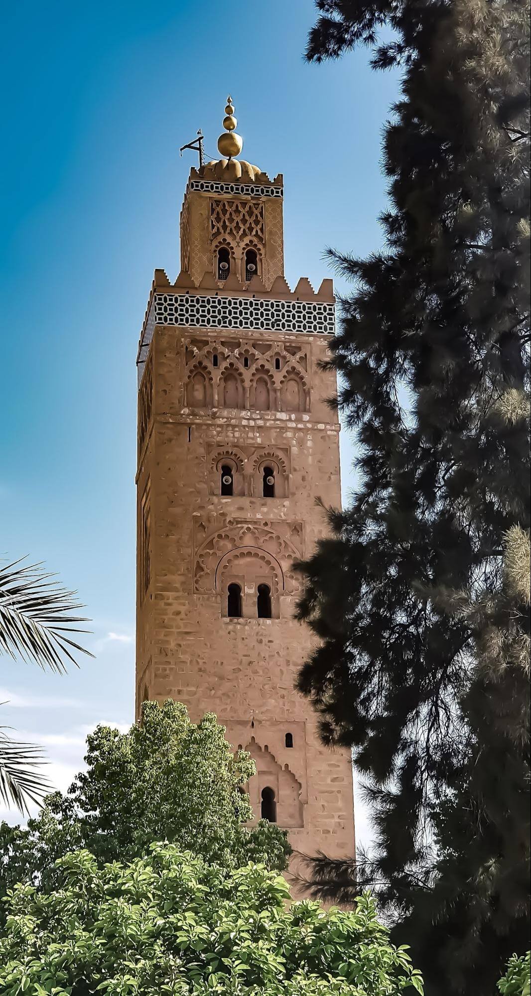 Visit Morocco: A Luxury Journey Through Imperial Cities, Atlas Peaks and Sahara Dunes 9 The historic Koutoubia Mosque minaret rising above lush green trees in Marrakech against a clear blue sky